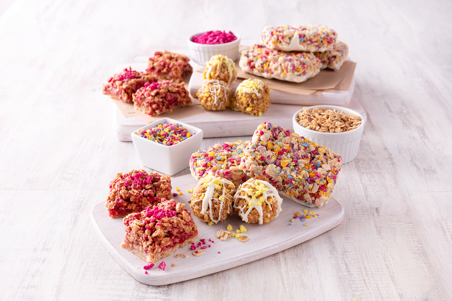 Assorted colorful treat bars, cookies, and toppings on wooden serving boards and bowls, with sprinkles, crushed cereal, and a bowl of rolled oats, on a white wooden surface.