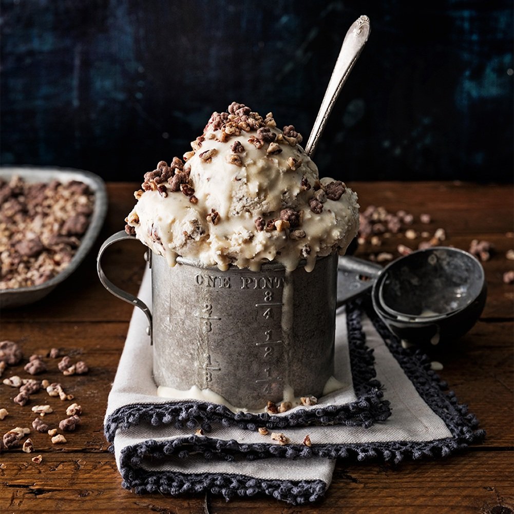 A metal measuring cup filled with homemade ice cream topped with chocolate and nut crumbles, placed on a cloth napkin on a wooden table, with a spoon inserted into the ice cream and a bowl of nut crumbles in the background.