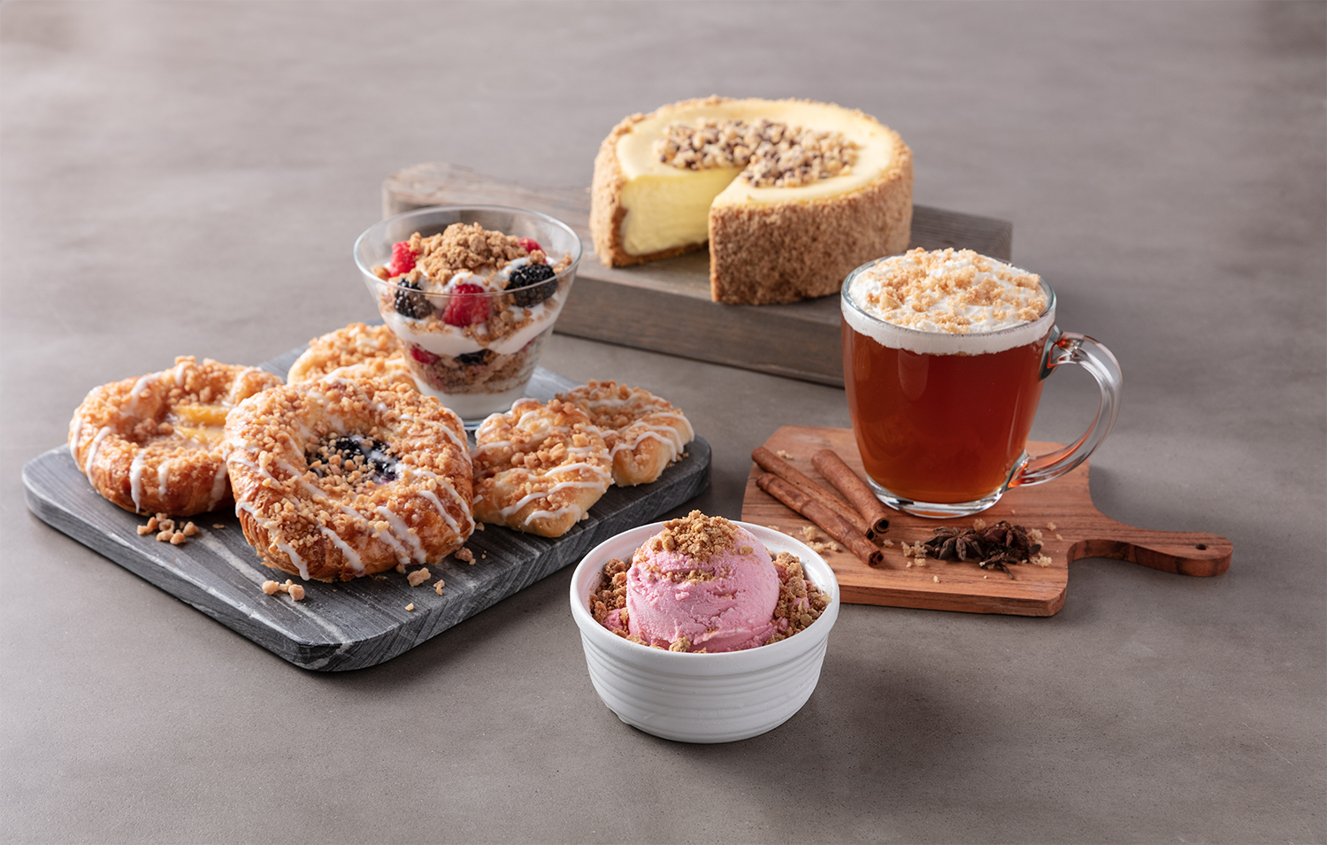 Assorted desserts including berry parfait, cheesecake, donuts, and pink ice cream, along with a cup of tea and cinnamon sticks.