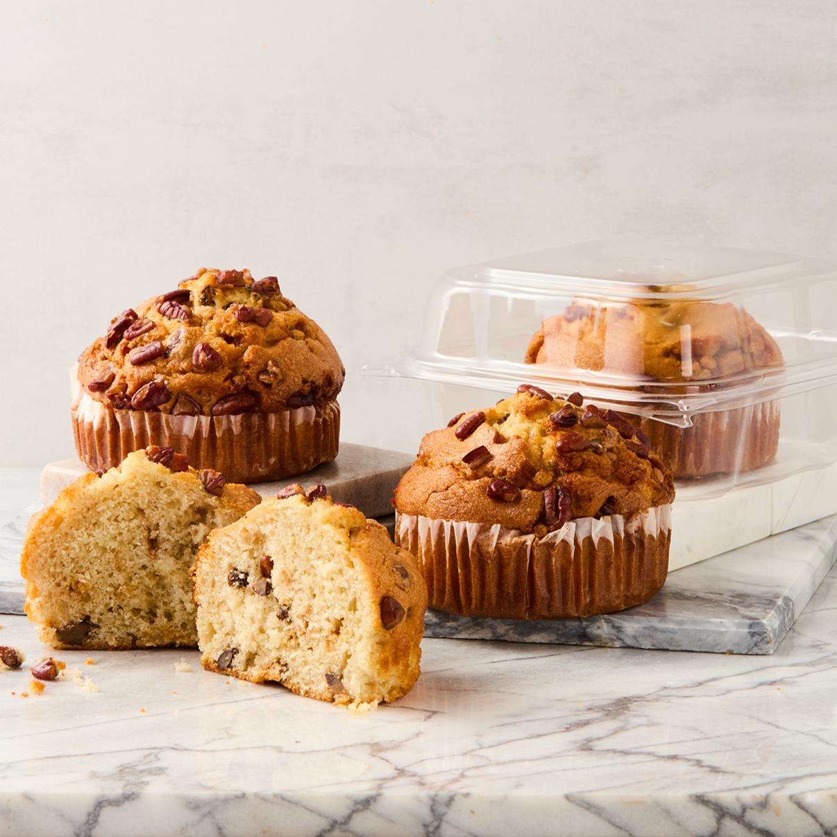 Several chocolate chip muffins, one cut in half, on a marble surface. Some muffins are in plastic containers, and one muffin is on a small grey marble square.