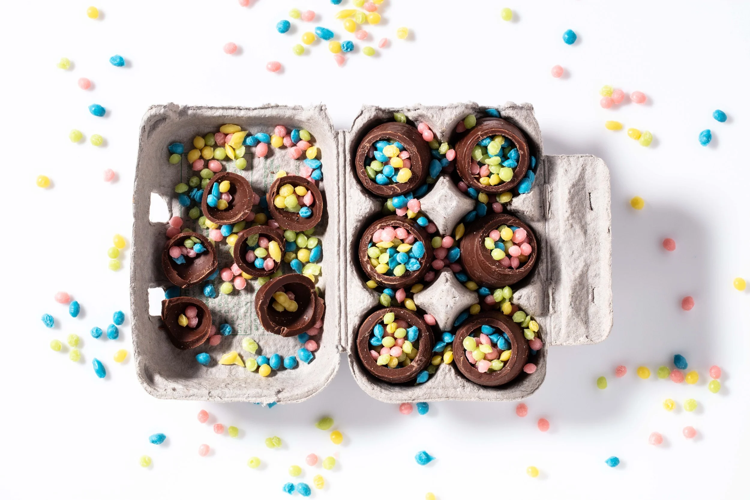 An open gray egg carton filled with chocolate eggs topped with pastel-colored sprinkles, with additional sprinkles scattered around on a white surface.