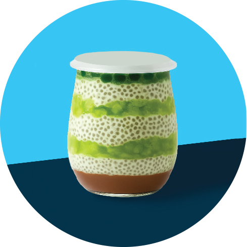 A layered dessert in a glass jar with green, white, or beige, and brown layers, topped with a white lid, against a two-tone blue background.