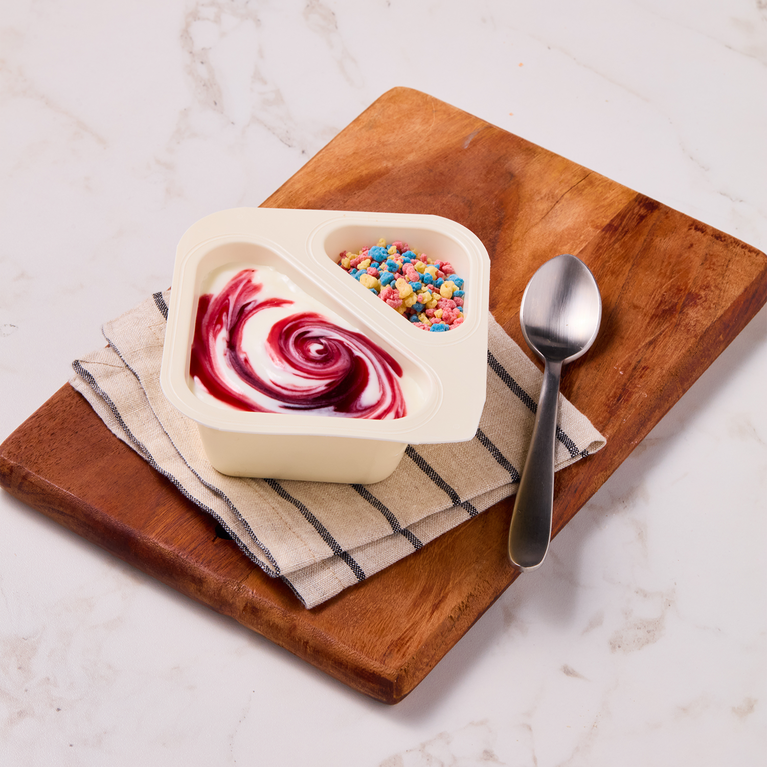 A container of yogurt with a swirl of berry sauce and colorful cereal pieces on a wooden cutting board with a spoon and a striped cloth napkin.
