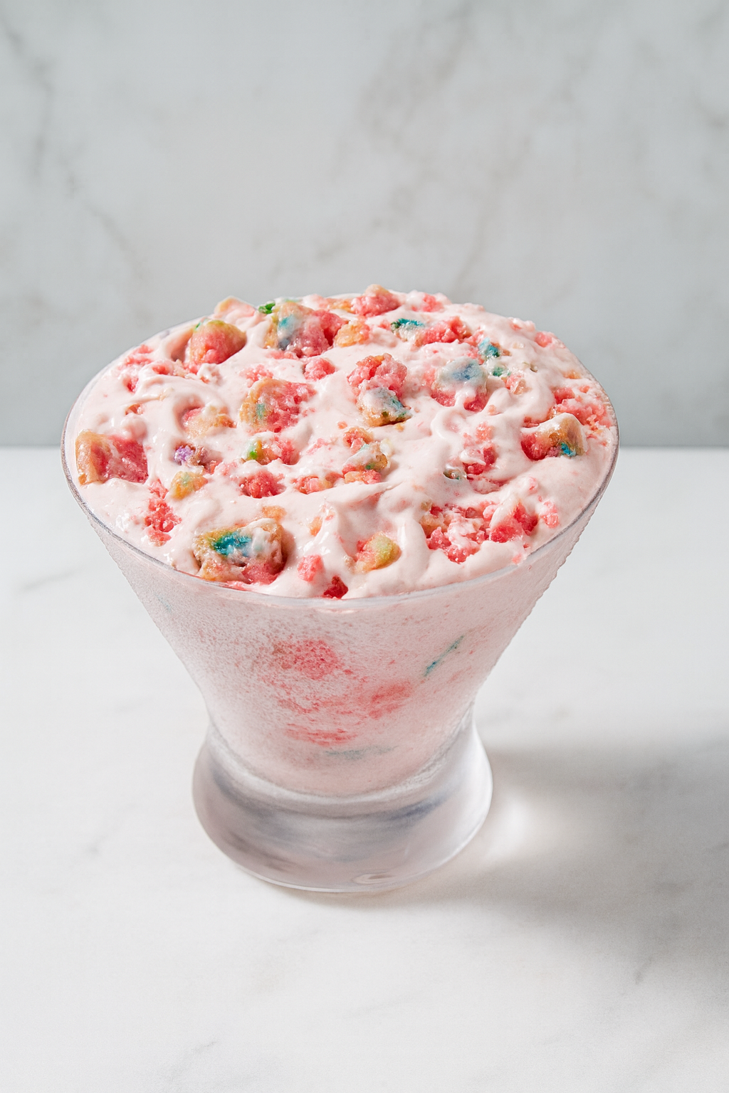 A glass of pink ice cream topped with colorful cereal pieces, served in a clear, footed glass bowl on a white surface with a light gray marble background.