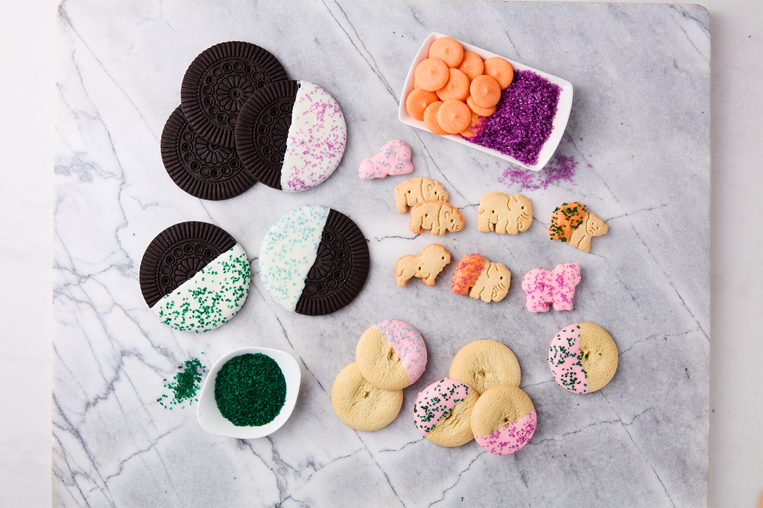 Easter cookies decorated with pink, blue, green, and purple sprinkles and local cookies in the shape of baby animals, with some pastel-colored candies and sugar sprinkles on a marble surface.