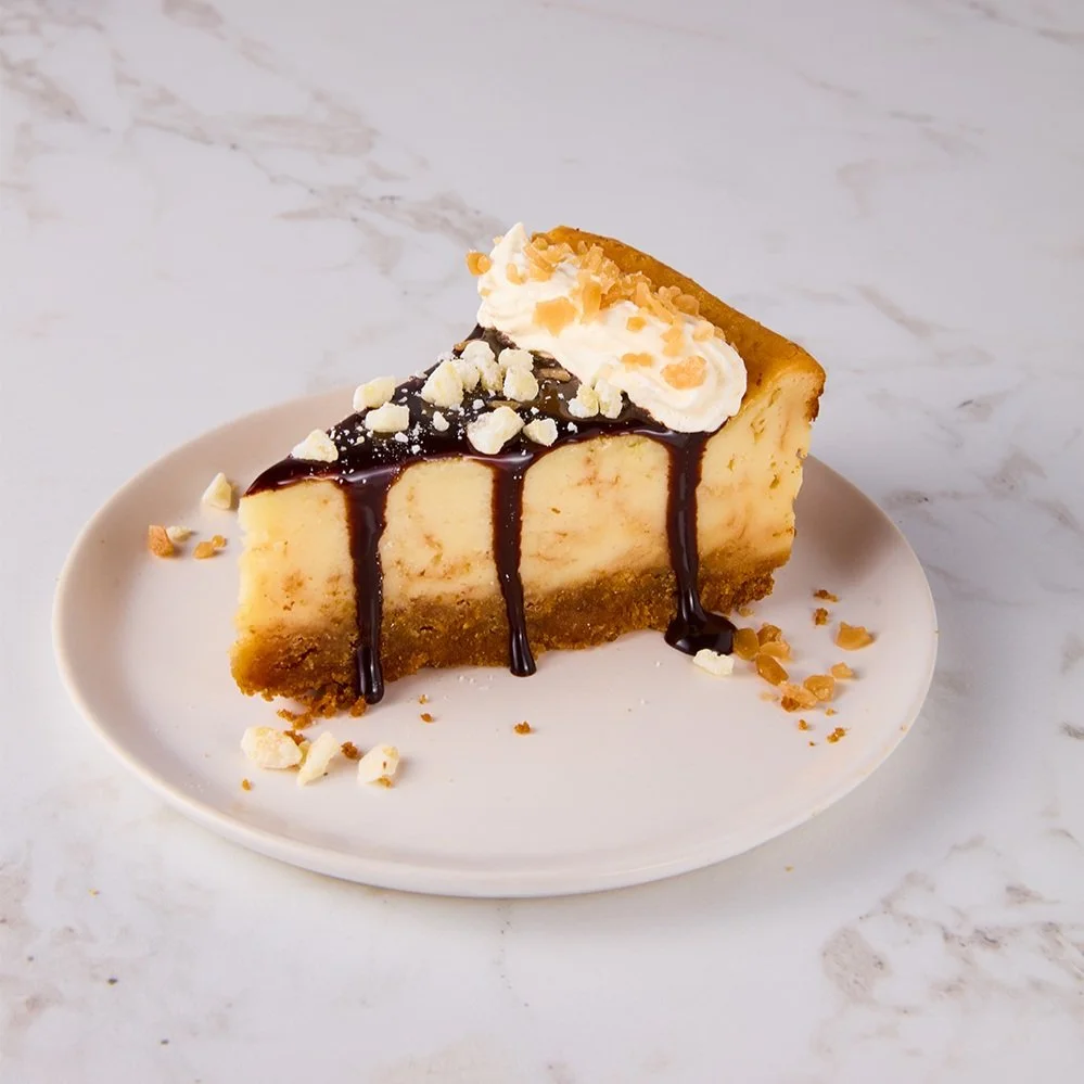 Slice of cheesecake with chocolate drizzle, whipped cream, and chopped nuts on a white plate
