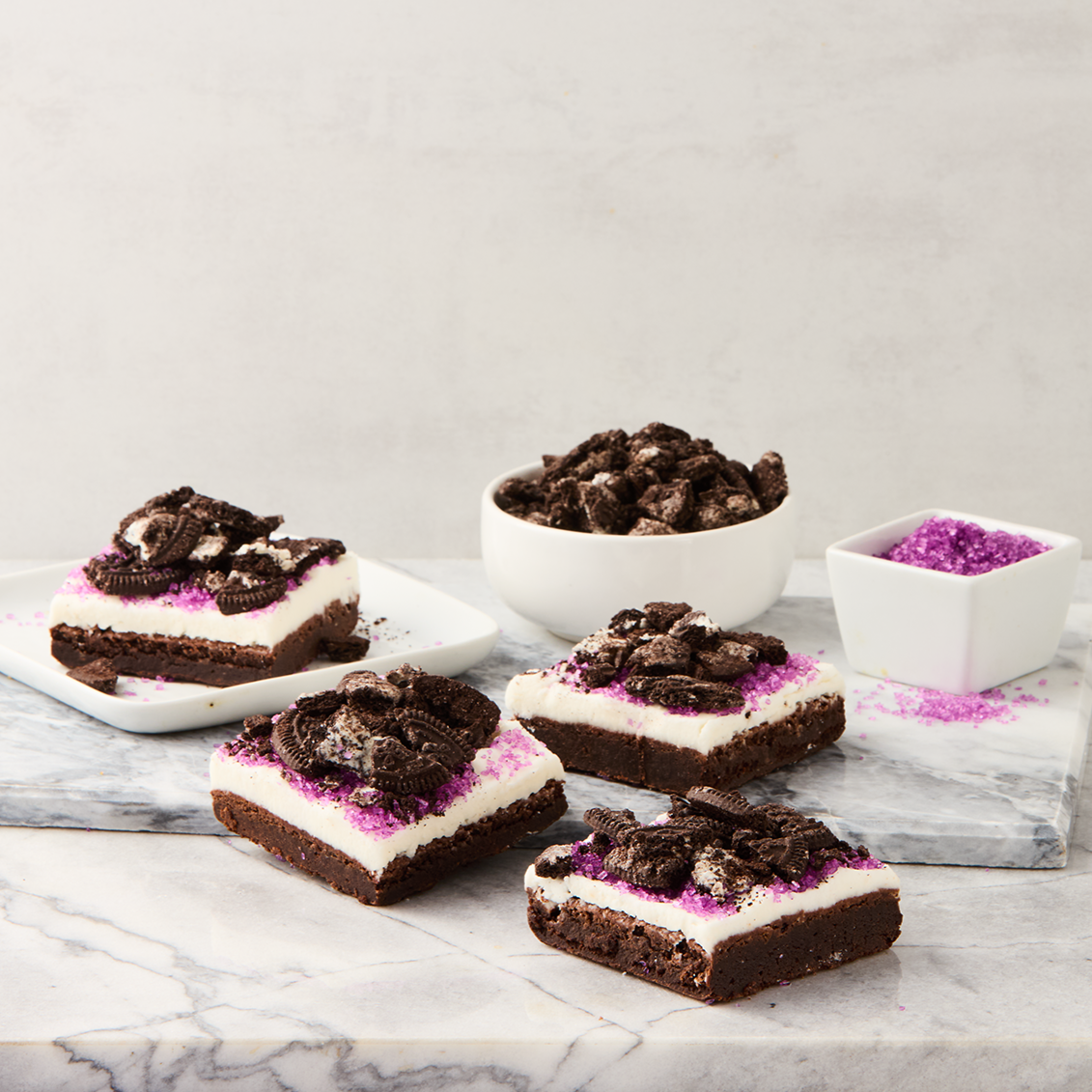 Chocolate Oreo layered dessert bars topped with crumbled Oreos and purple sprinkles, with bowls of crushed Oreos and purple sugar on a marble surface.