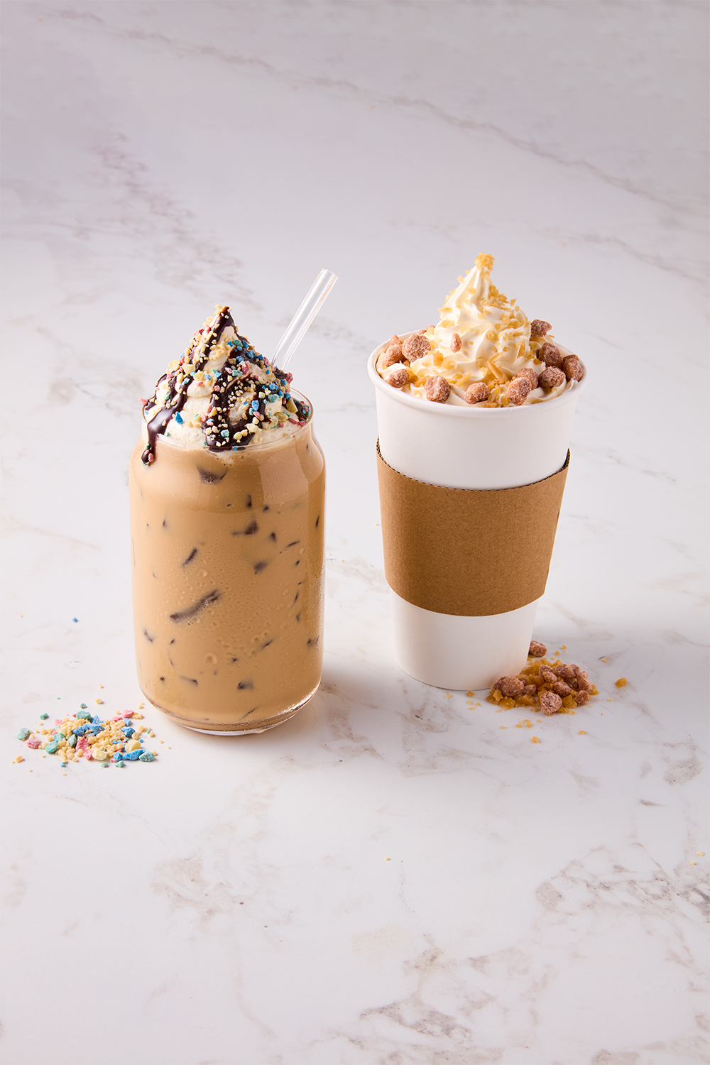 A mason jar filled with iced coffee topped with whipped cream, chocolate syrup, sprinkles, and a clear straw, and a white paper coffee cup with a whipped cream topping, cereal pieces, and a cardboard sleeve, sitting on a white marble surface.