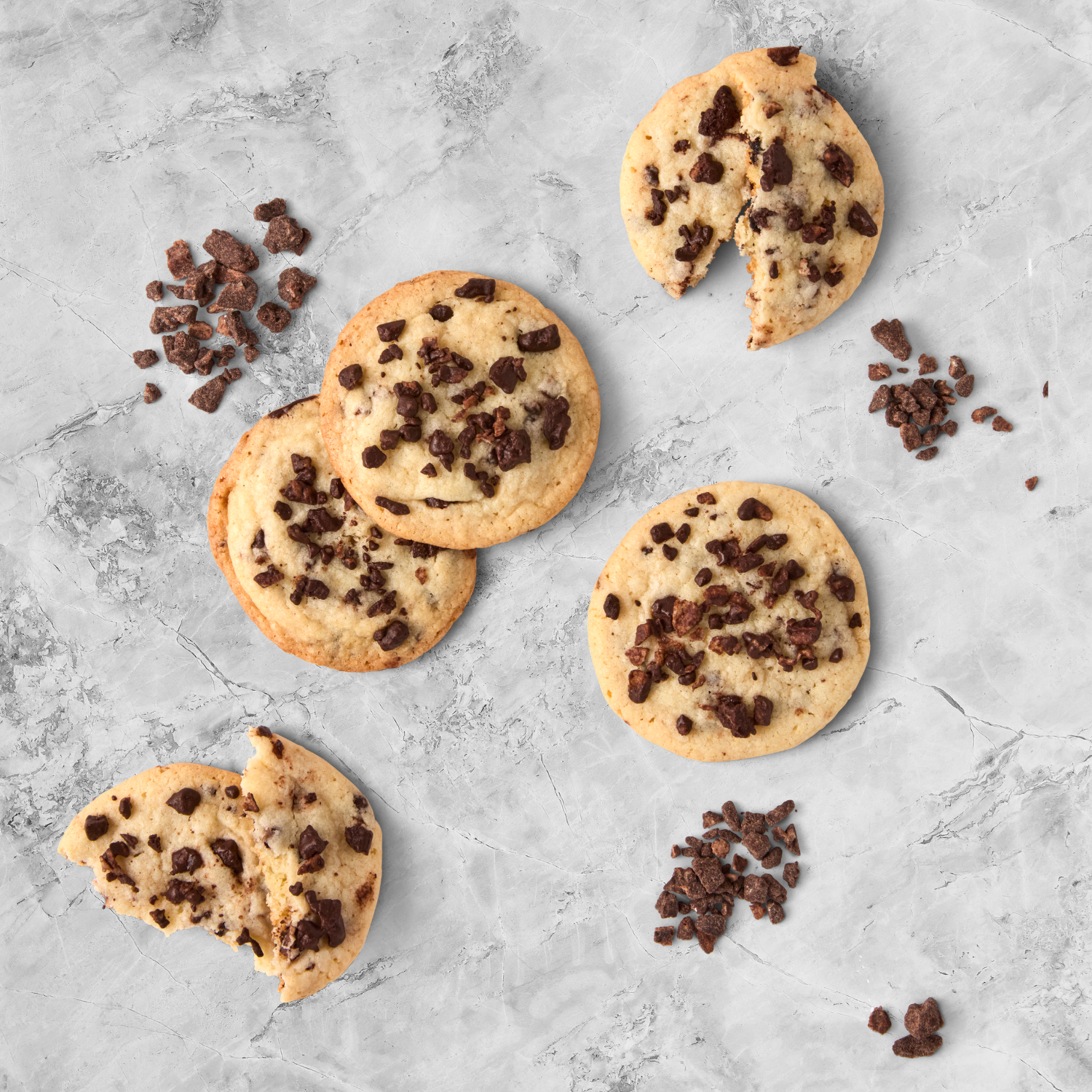 Chocolate chip cookies on a gray marble surface, with some broken pieces and chocolate crumbles scattered around.