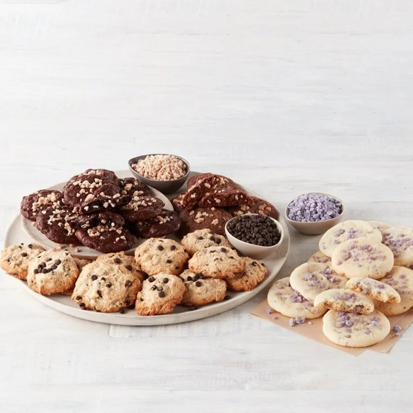 Assorted cookies, including chocolate chip, oatmeal, and decorated sugar cookies, with small bowls of toppings or sprinkles on a white surface.
