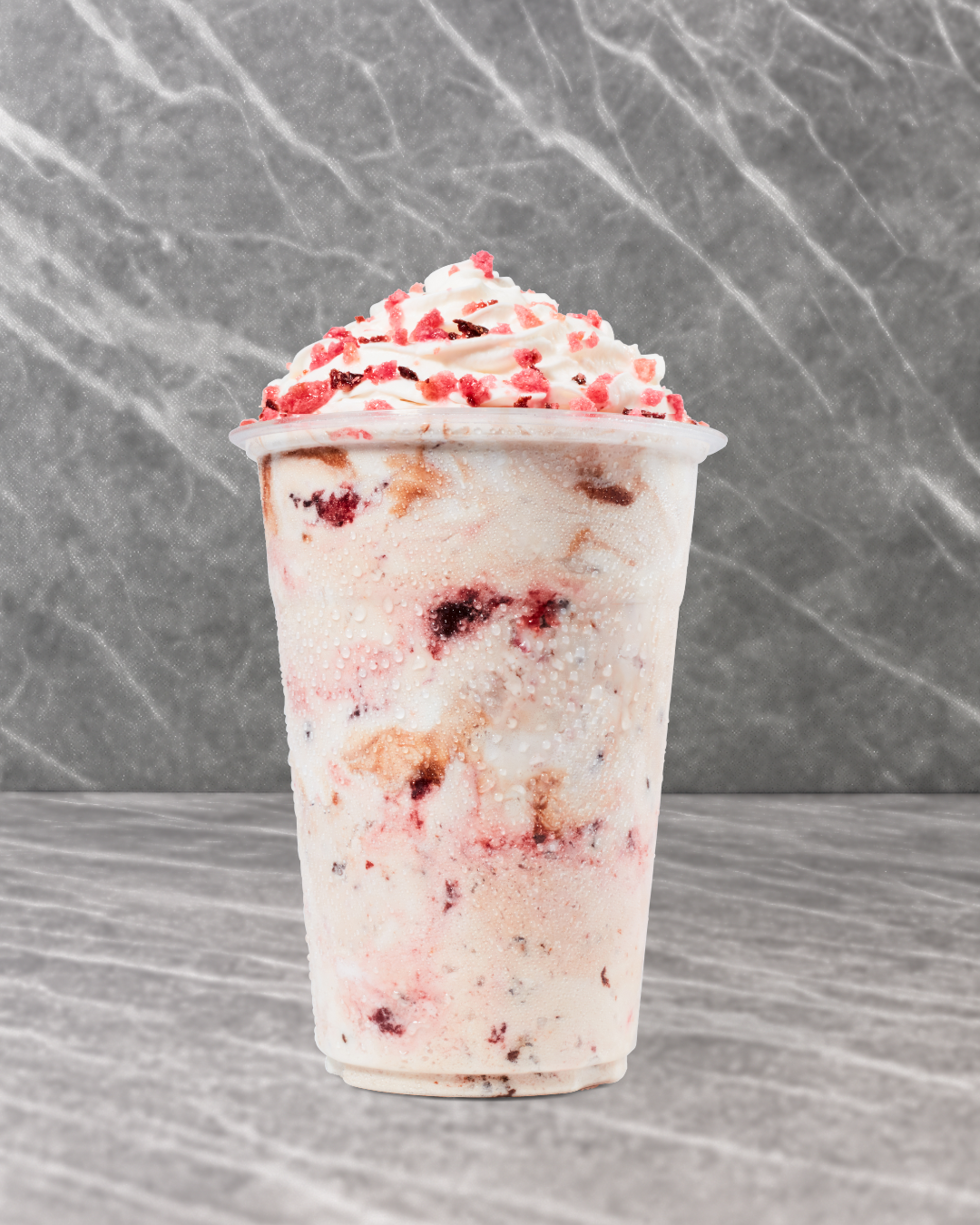 A cold beverage in a clear plastic cup topped with whipped cream and pink sprinkles, with visible chunks of berries and ice