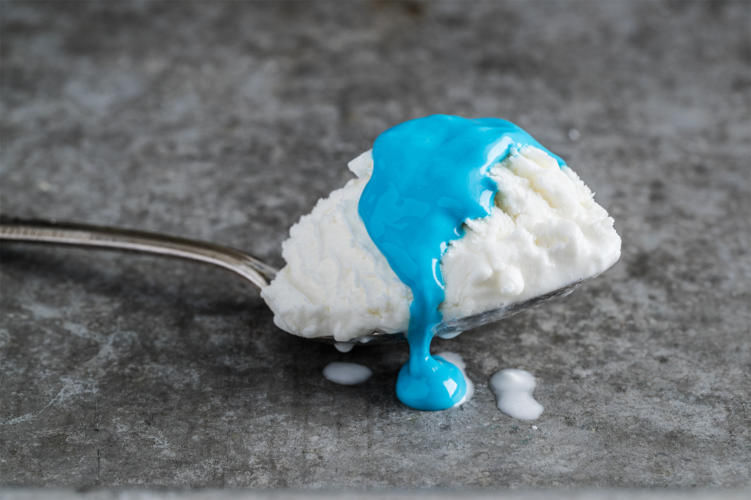 Scoop of vanilla ice cream with blue syrup drizzling over it, on a gray textured surface.