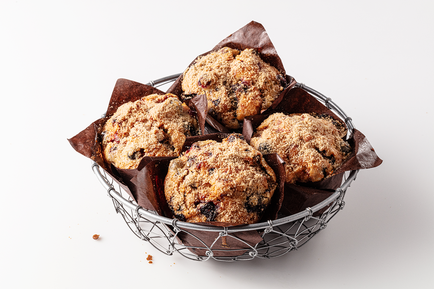 Four blueberry muffins with streusel topping in a small wire basket lined with brown paper, on a white surface.