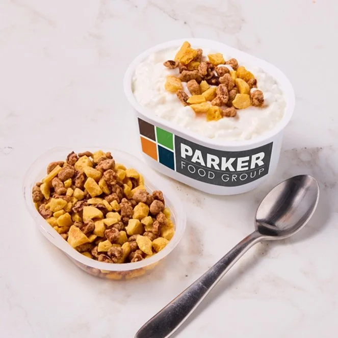 Two bowls of yogurt topped with granola and fruit, one labeled PARKER FOOD GROUP, with a metal spoon placed on a white surface.