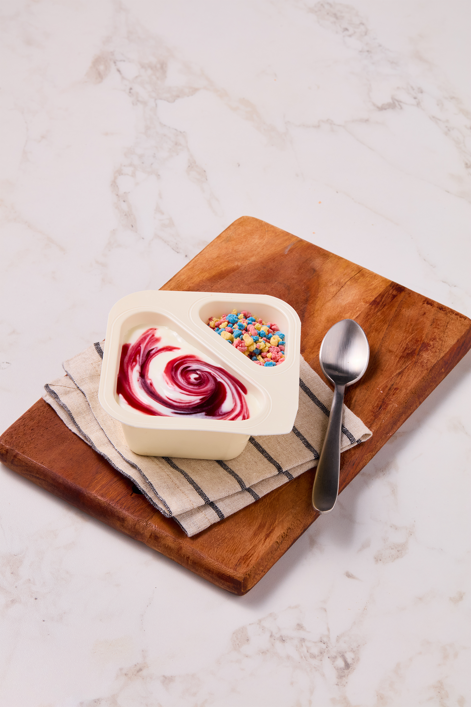 A bowl of yogurt with a swirl of raspberry sauce and colorful cereal pieces on a wooden board with a spoon.