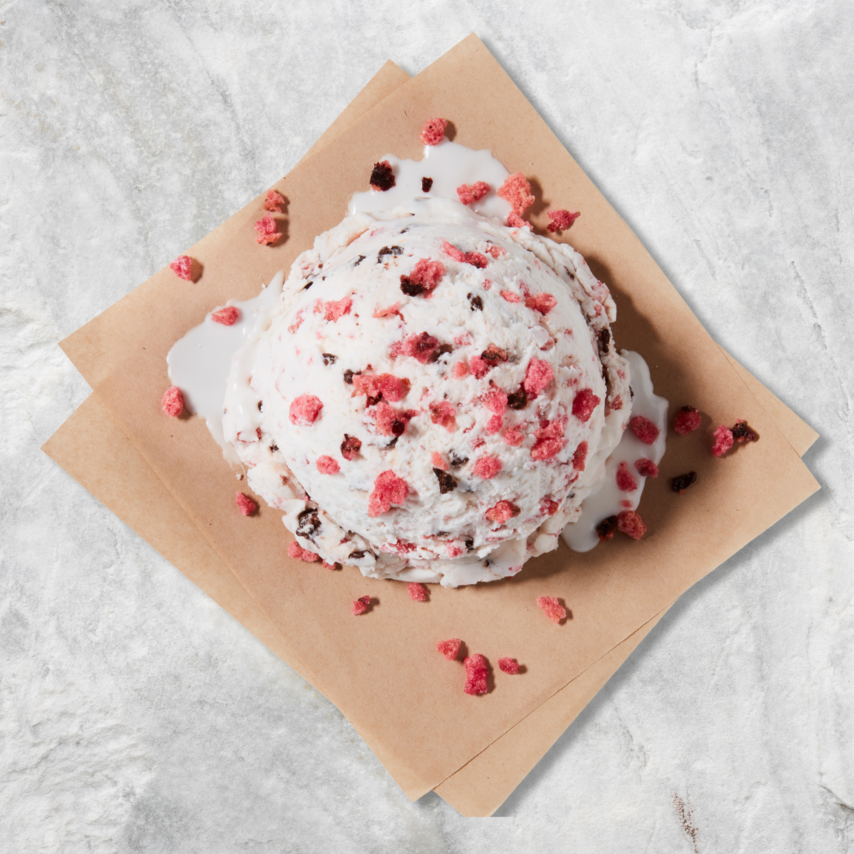 A scoop of ice cream with pink and brown berry pieces, served on a piece of brown parchment paper.