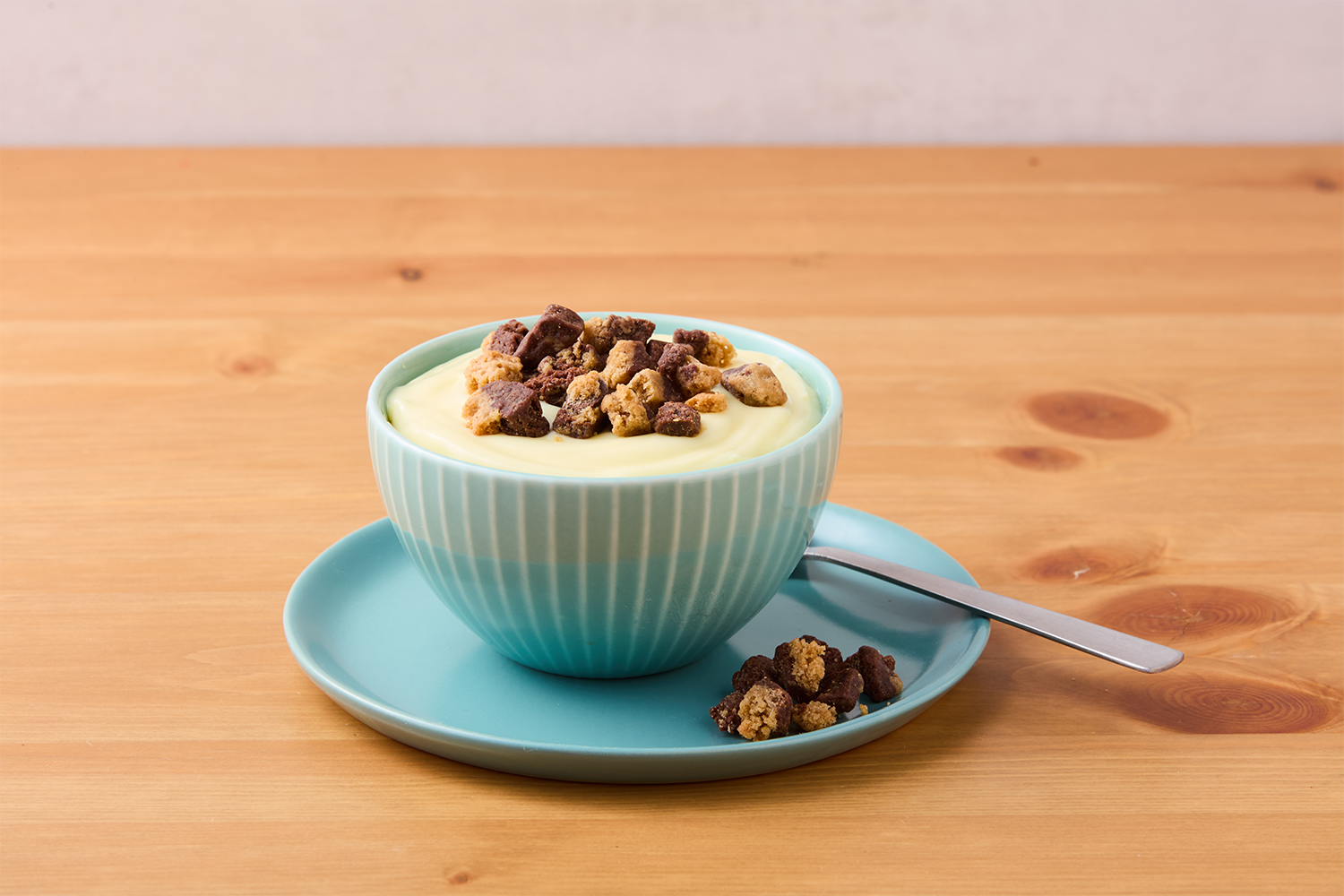A bowl of creamy custard topped with chocolate and vanilla cookie crumbles, sitting on a matching blue plate with a spoon, on a wooden table.
