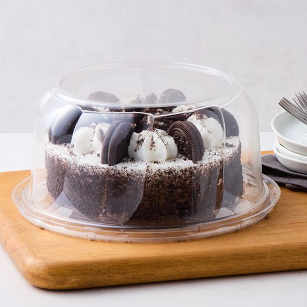 Chocolate cake topped with Oreo cookies and white whipped cream, covered with a clear plastic cake dome on a wooden cutting board.