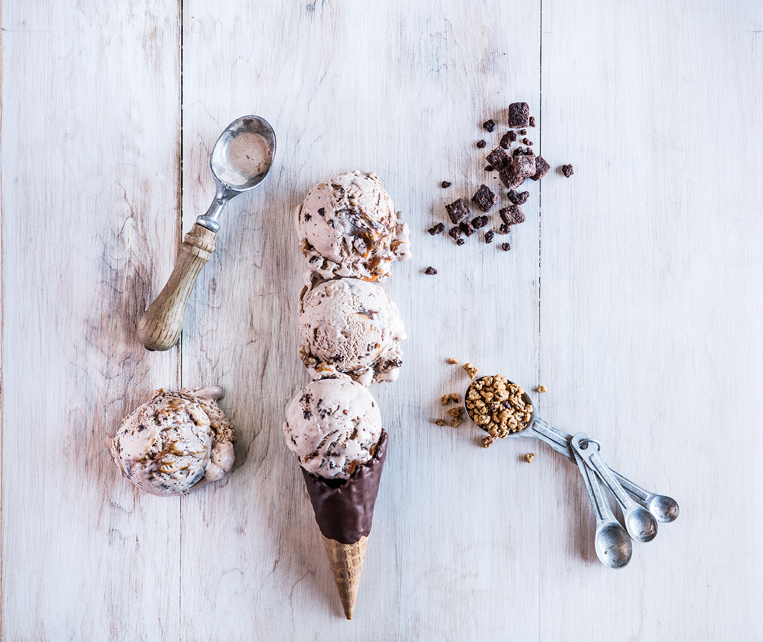 Three scoops of ice cream with chocolate chunks, a smaller scoop of ice cream, a chocolate-coated ice cream cone, a tablespoon of caramelized nuts, a scoop of nuts, and scattered chocolate cubes on a white wooden surface.