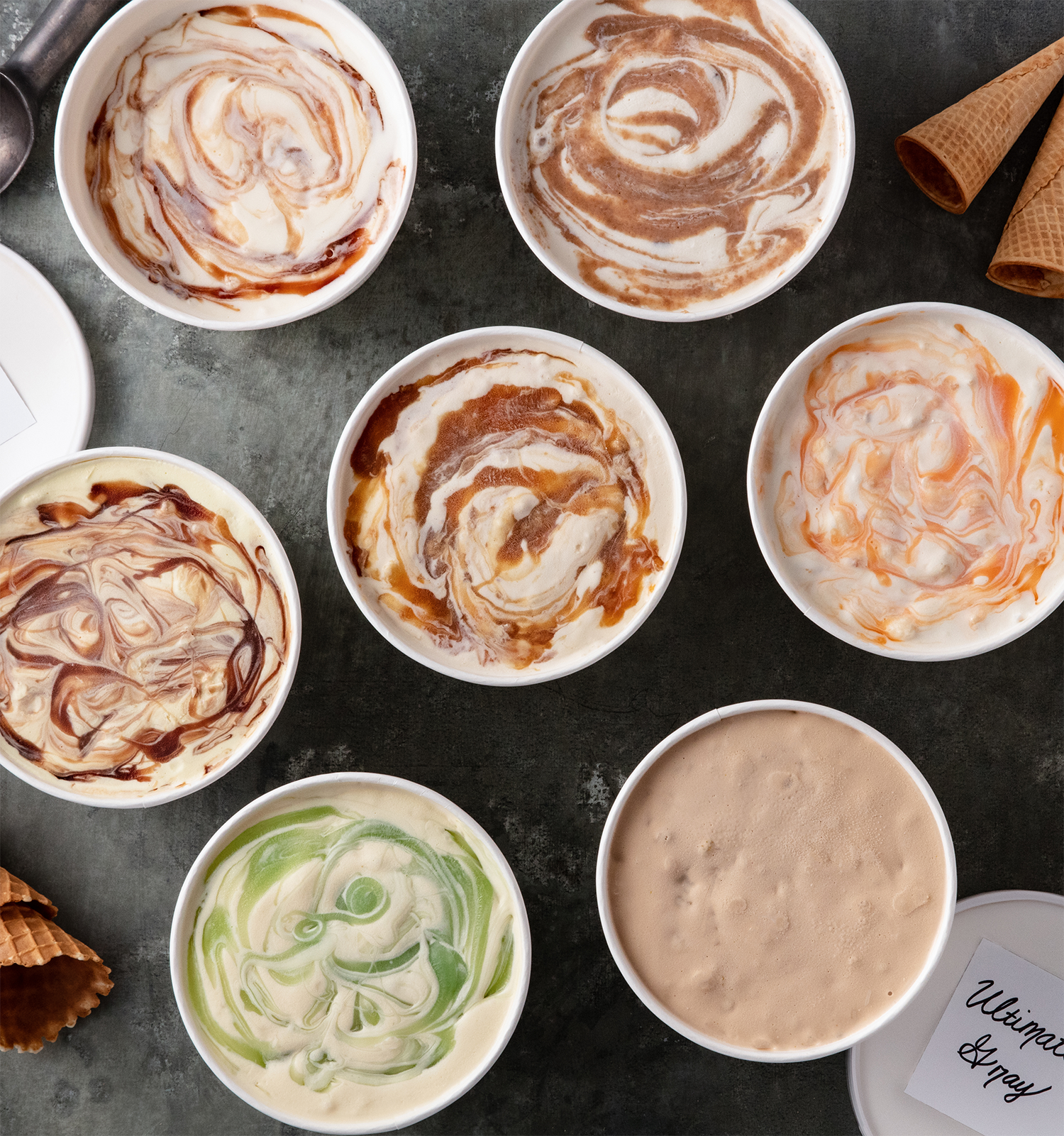 Various flavors of ice cream with swirls of caramel, chocolate, and fruit in bowls on a dark surface, with waffle cones in the background.