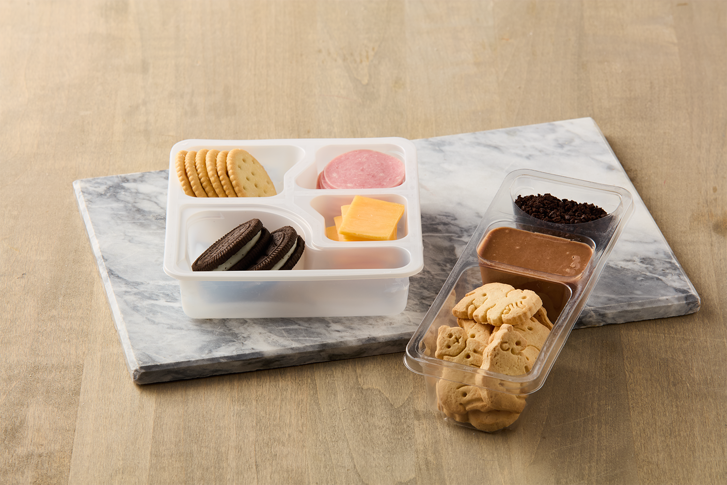 Assorted cookies, crackers, cheese, ham, and chocolate sauce in a plastic container on a marble cutting board.