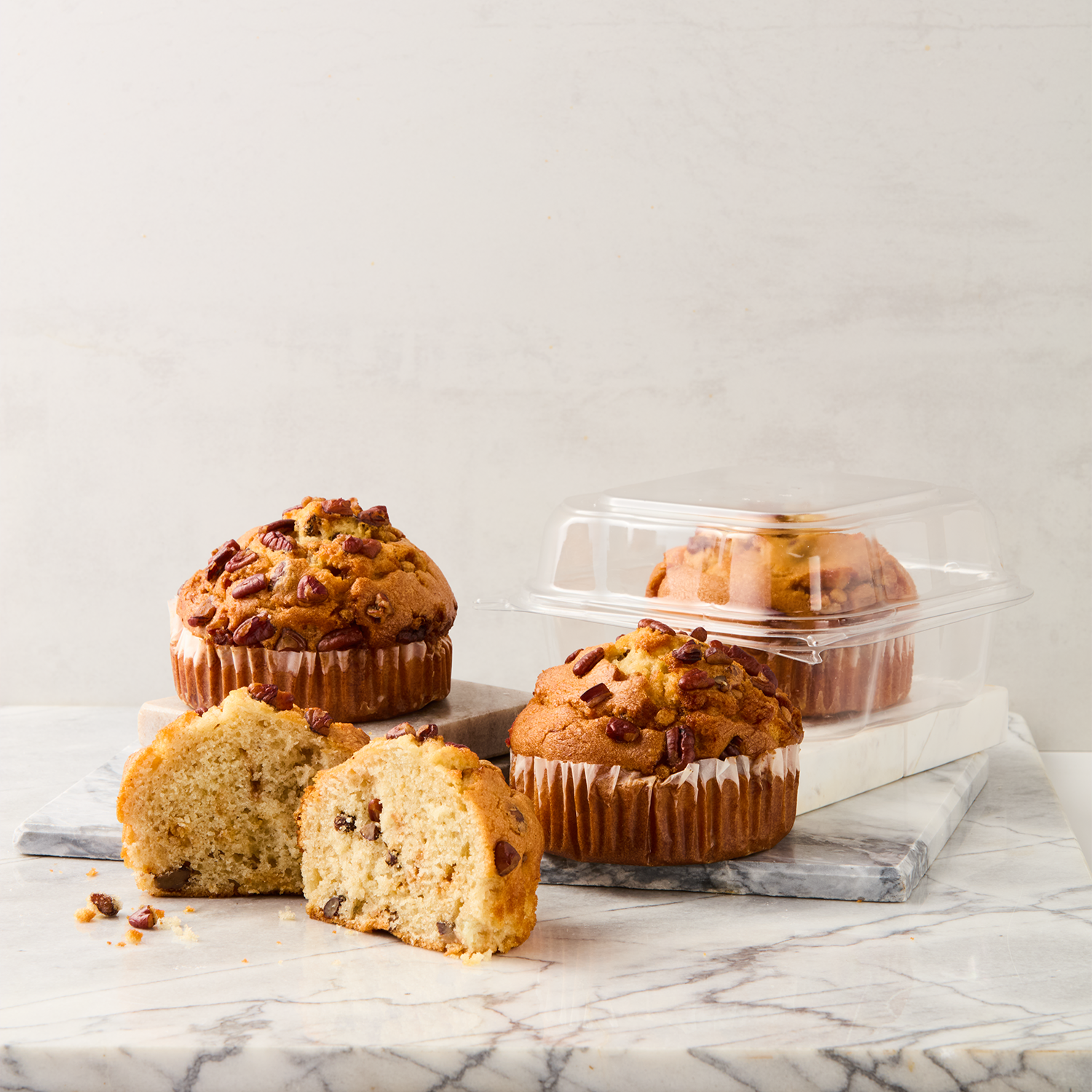 Three chocolate chip muffins on a marble surface, one cut in half revealing the inside with chocolate chips, with two muffins in plastic containers behind.