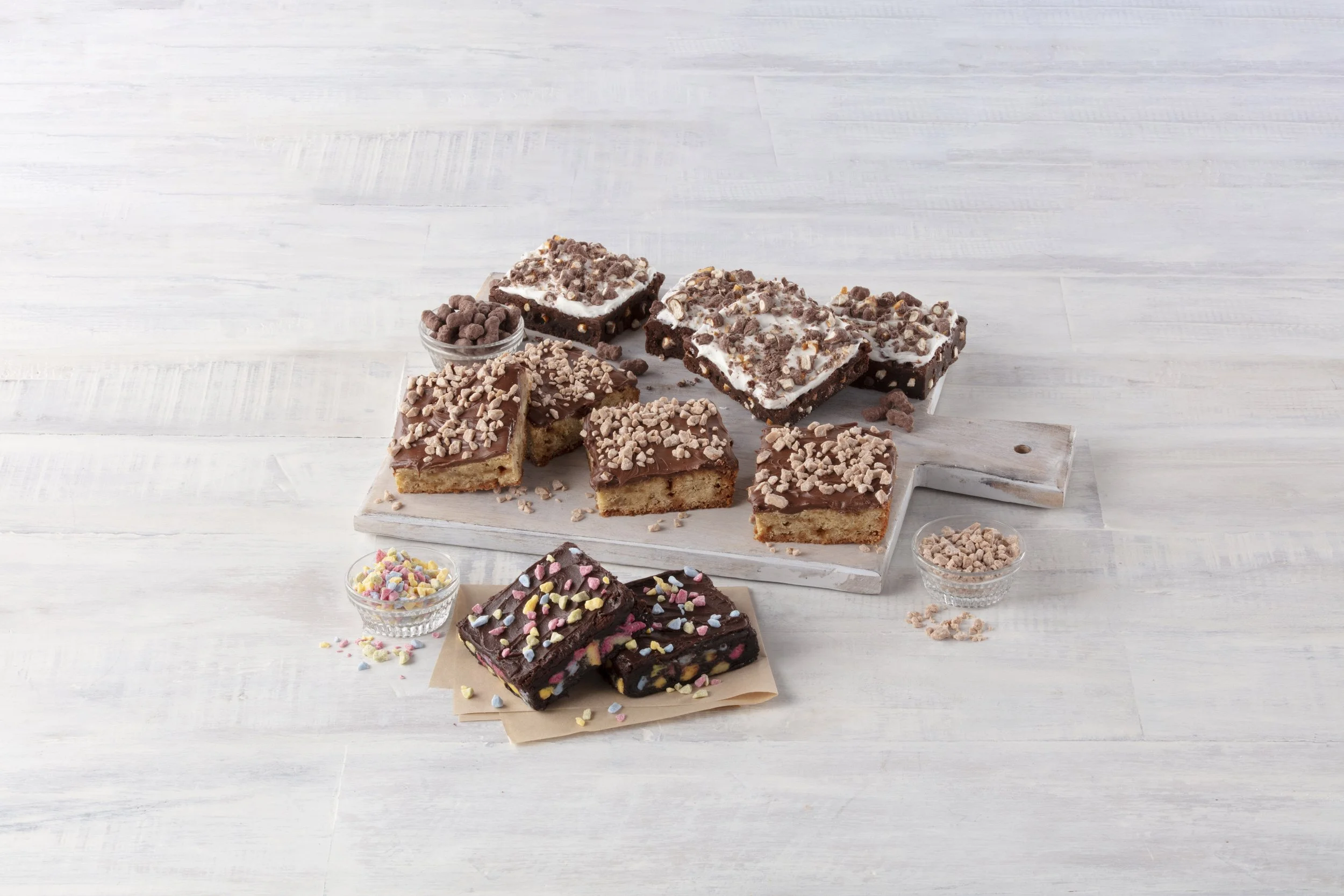 Assorted brownies and blondies with sprinkles, chocolate chips, and other toppings on a wooden serving board, with bowls of toppings nearby on a white wooden surface.