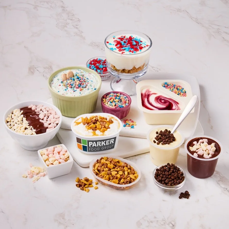 Assorted containers of frozen yogurt toppings, including sprinkles, chocolate chips, fruit pieces, and flavored syrups, arranged on a white marble surface.