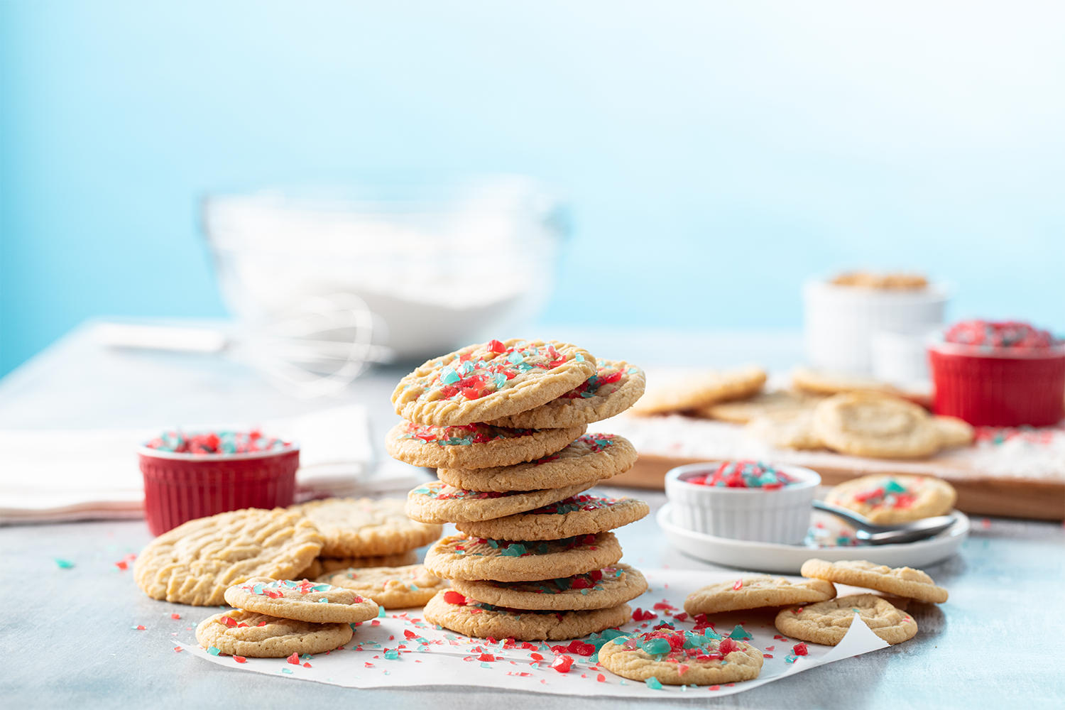 Stack of decorated sugar cookies with red and blue sprinkles on a white parchment paper, with additional cookies and sprinkles around, on a light blue surface, set against a blurred background of baking ingredients and a whisk.