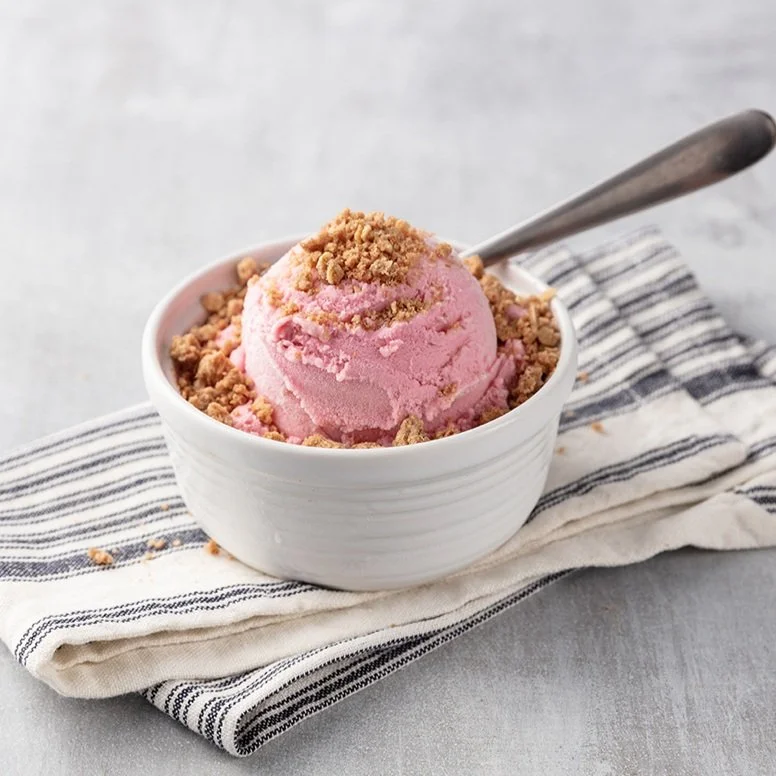 A white bowl of pink ice cream topped with crumbled cookies and a spoon, placed on a striped cloth napkin on a gray surface.