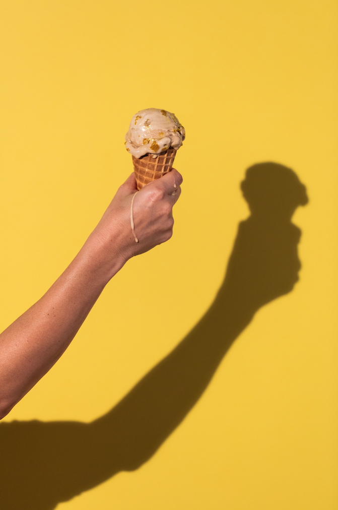 Hand holding an ice cream cone with a yellow background and a shadow of the hand and ice cream cone.