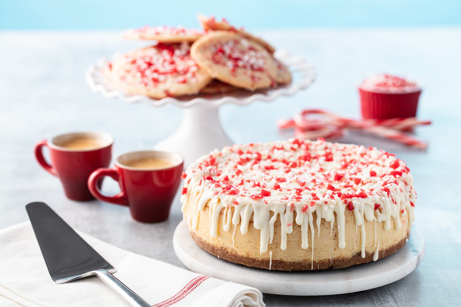 Festive holiday cookies and cheesecake with white chocolate drizzle and peppermint sprinkles on a marble cake stand, with two cups of coffee and red and white candy canes on a light-colored table.