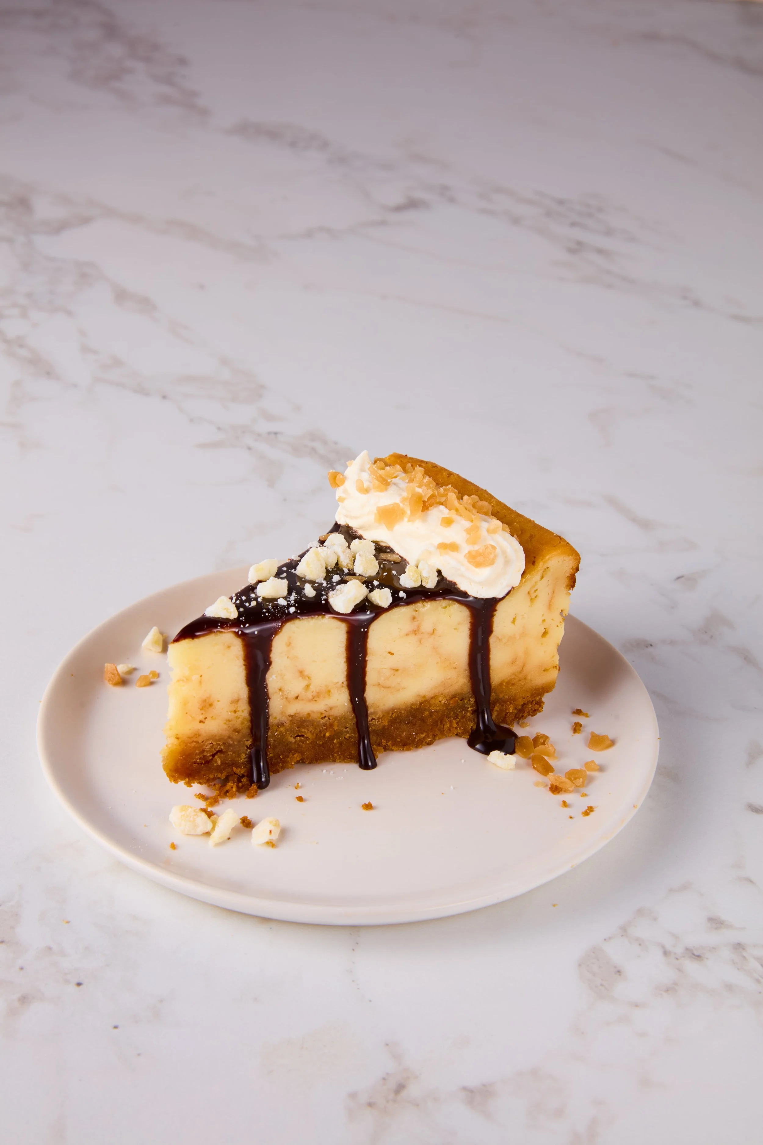 Slice of cheesecake topped with whipped cream, chocolate sauce, and chopped nuts on a white plate.