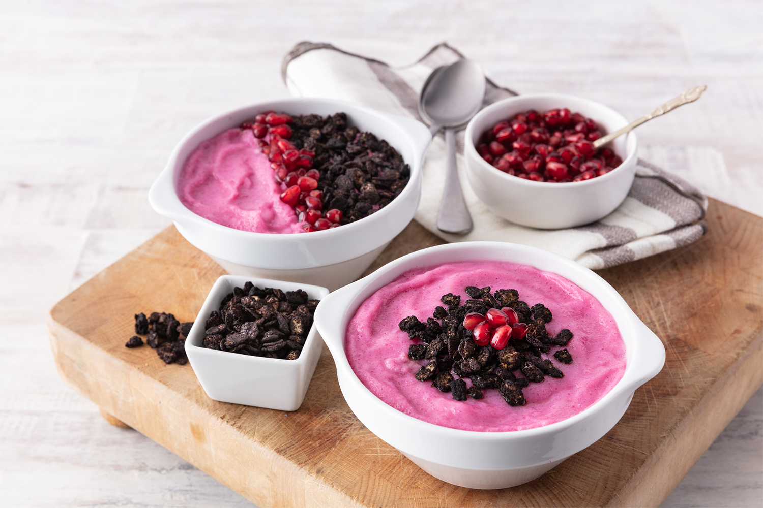 Two white bowls of pink berry dessert topped with pomegranate seeds and crumbled dark chocolate, surrounded by additional dark chocolate and pomegranate seeds, on a wooden cutting board with a striped napkin and spoons.