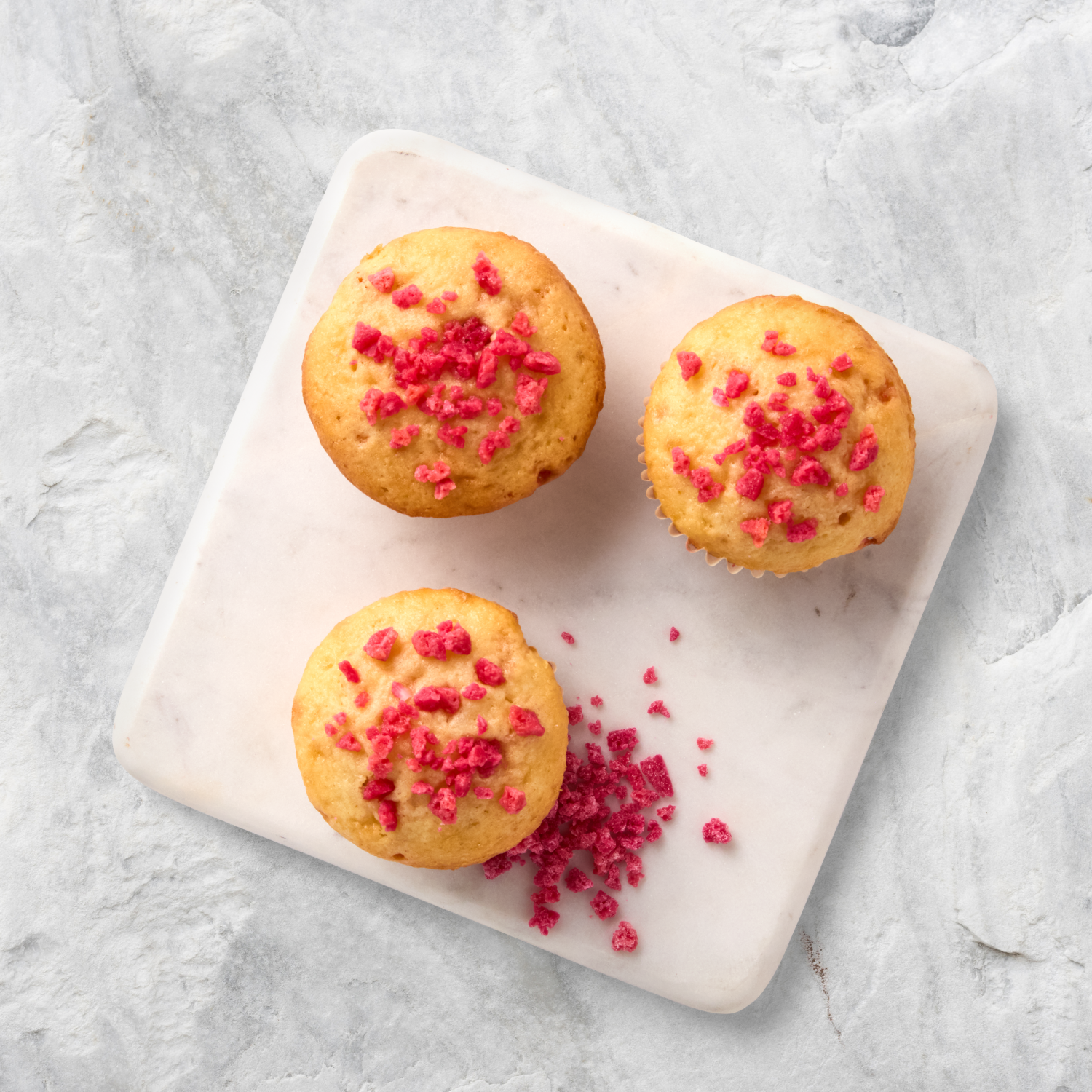 Three vanilla cupcakes with pink sprinkles on a white marble tray.
