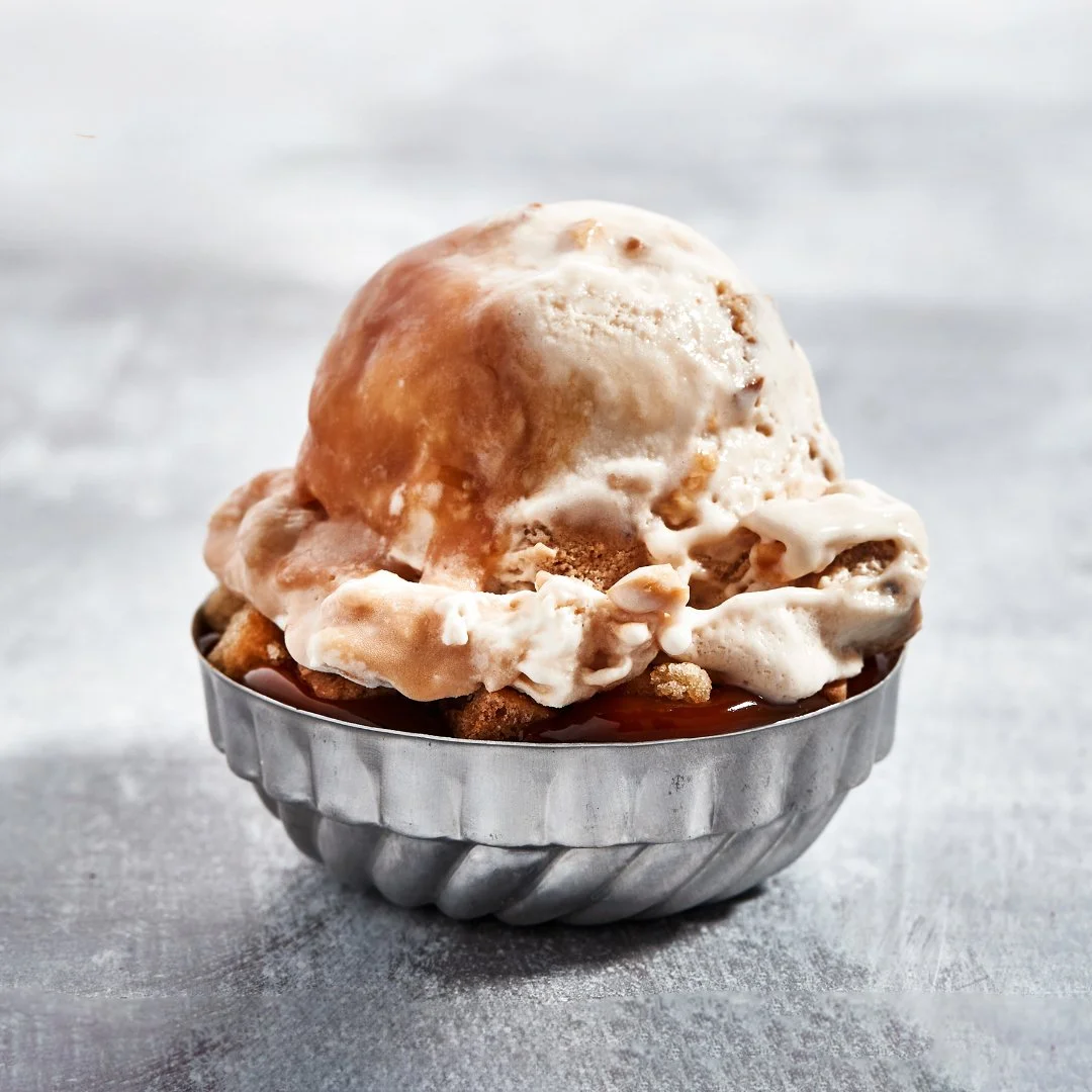 A scoop of vanilla ice cream in a small metal dish with toppings and cookie crumbs underneath.