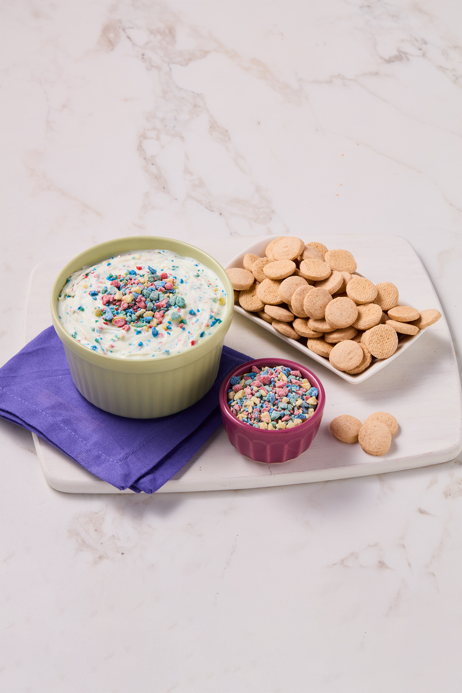 A bowl of cake batter topped with colorful sprinkles, a small pink cup of more sprinkles, and a plate of round cookies on a white marble countertop with a purple napkin underneath the bowl.