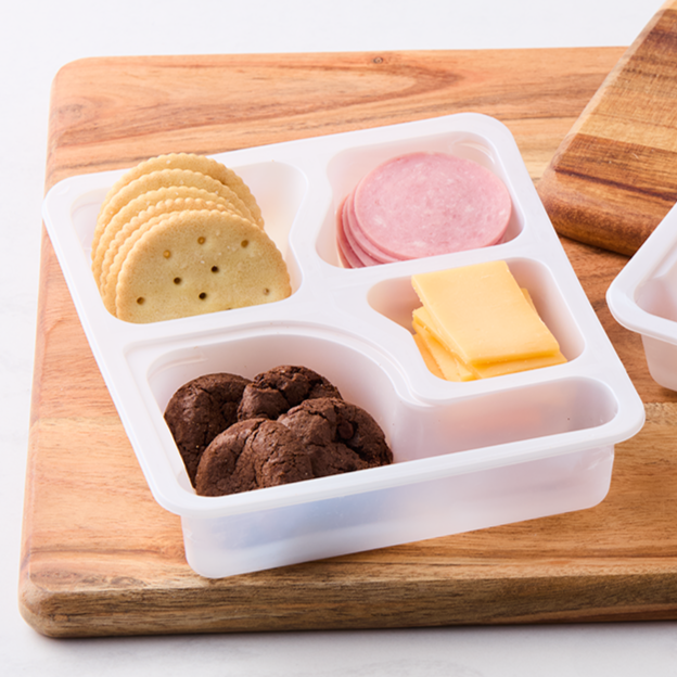 Sectioned plastic food container with round crackers, slices of ham, slices of cheddar cheese, and chocolate cookies on a wooden cutting board.