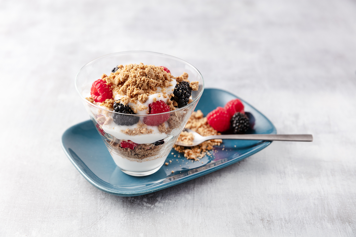 A dessert in a glass with layers of crushed cookies, fresh raspberries and blackberries, whipped cream, and crumbled toppings, served on a blue plate with a spoon, on a light surface.