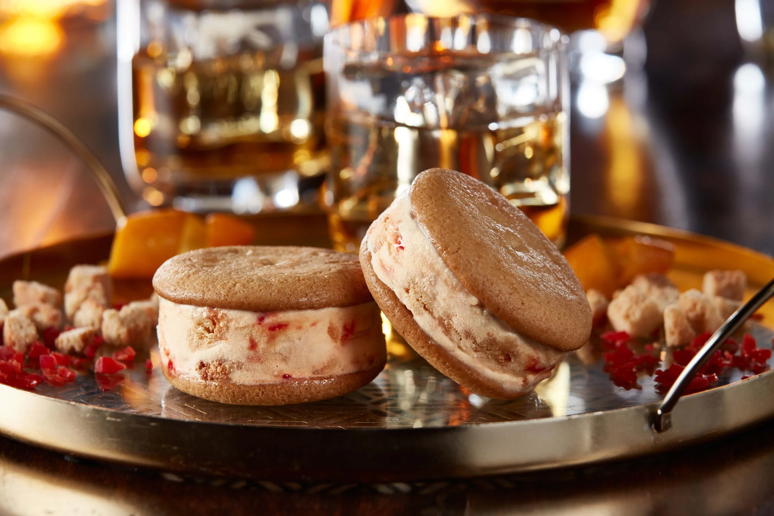 Two ice cream sandwiches with vanilla ice cream and red fruit bits, along with crumbles and a sip of whiskey in the background on a reflective gold tray.