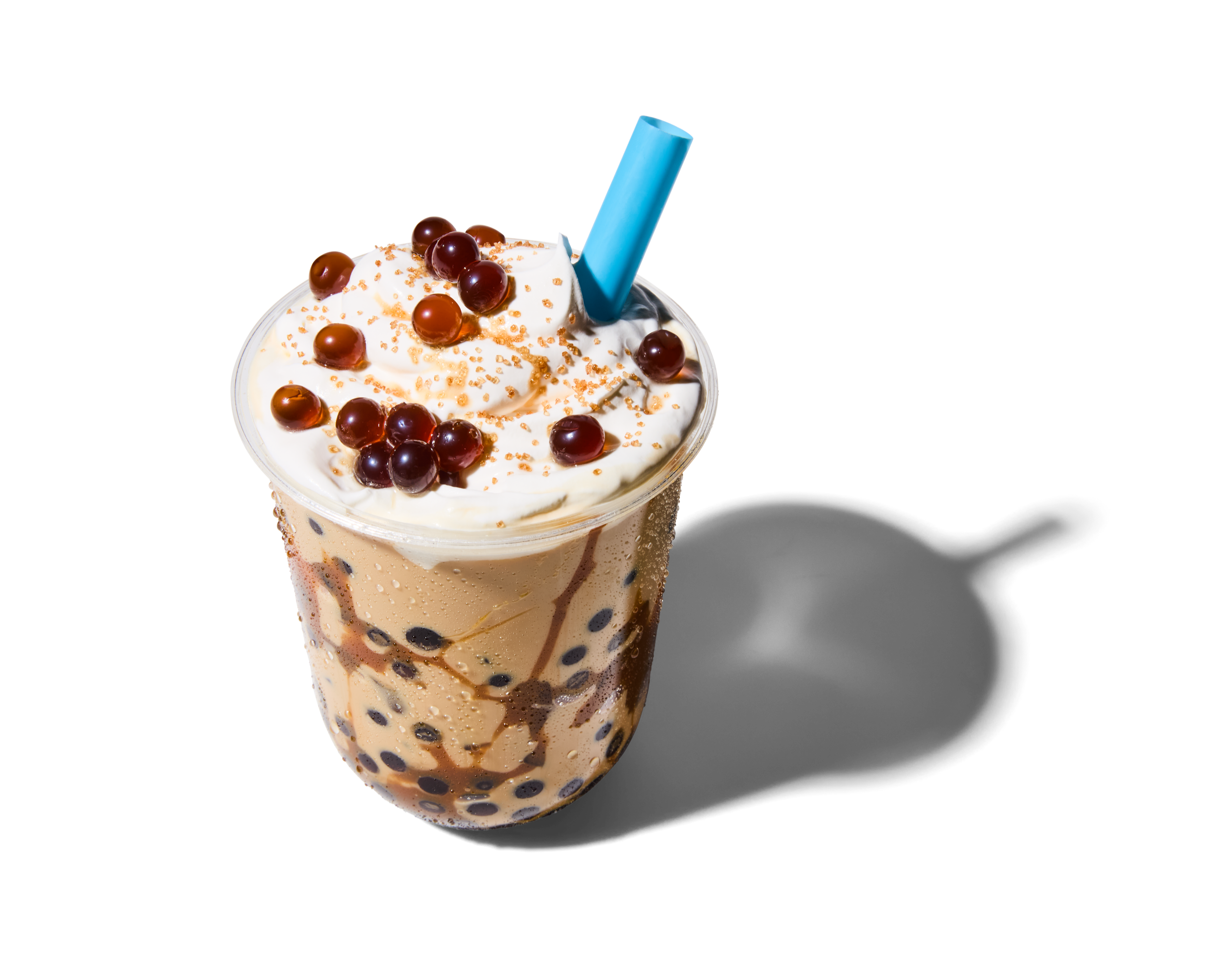 A milkshake with whipped cream, chocolate pearls, and sprinkles in a clear glass with a blue straw.