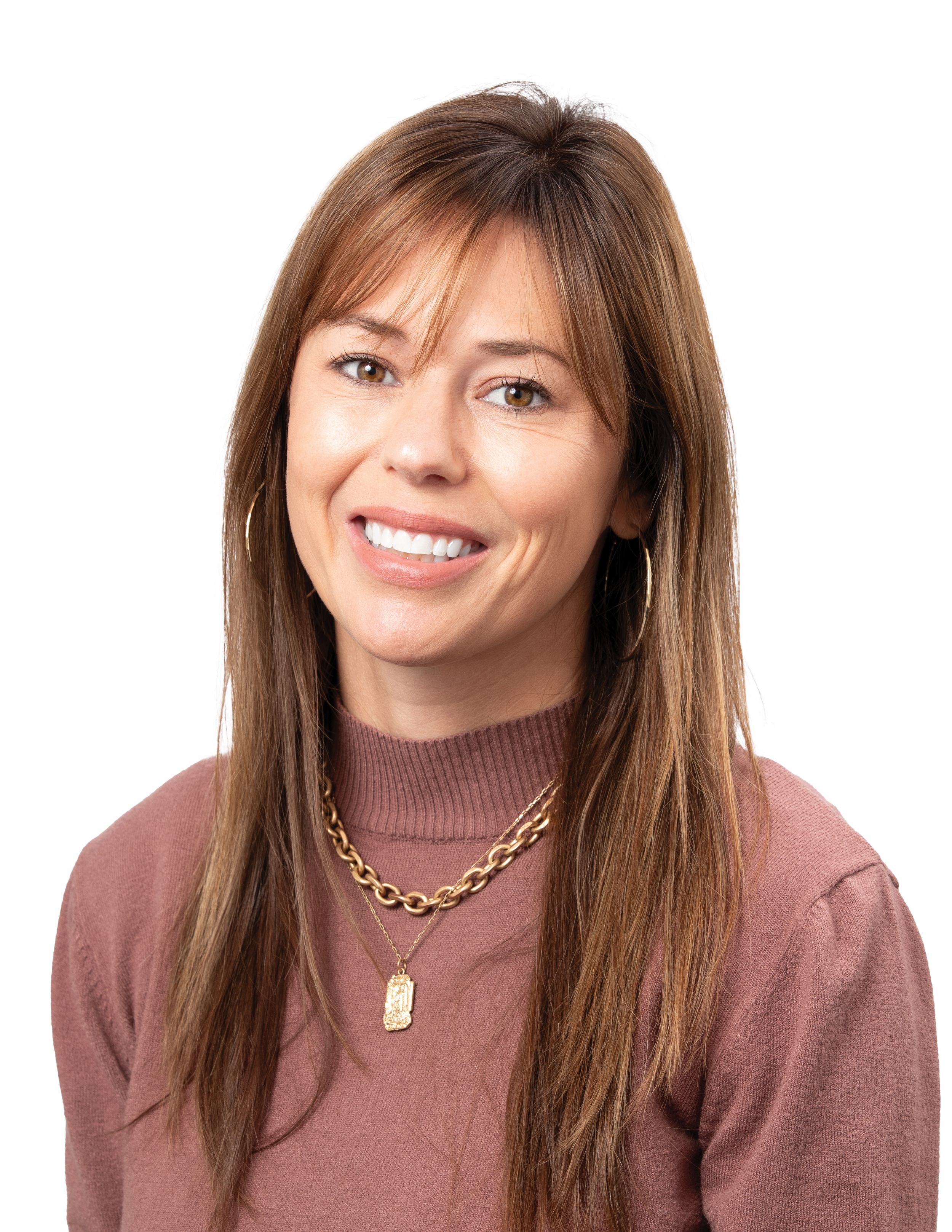 Close-up of a woman with brown hair, wearing a mauve sweater, gold chain necklace, and gold pendant, smiling with white teeth.