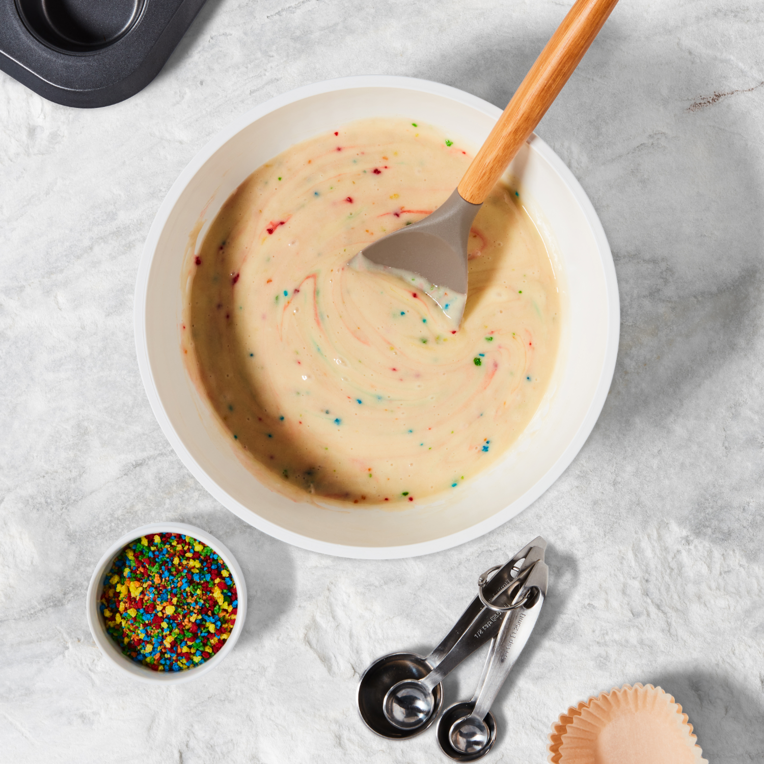 A bowl of rainbow sprinkles ice cream with a scooper, a small bowl of colorful sprinkles, metal measuring spoons, and a cupcake liner on a gray marble surface.