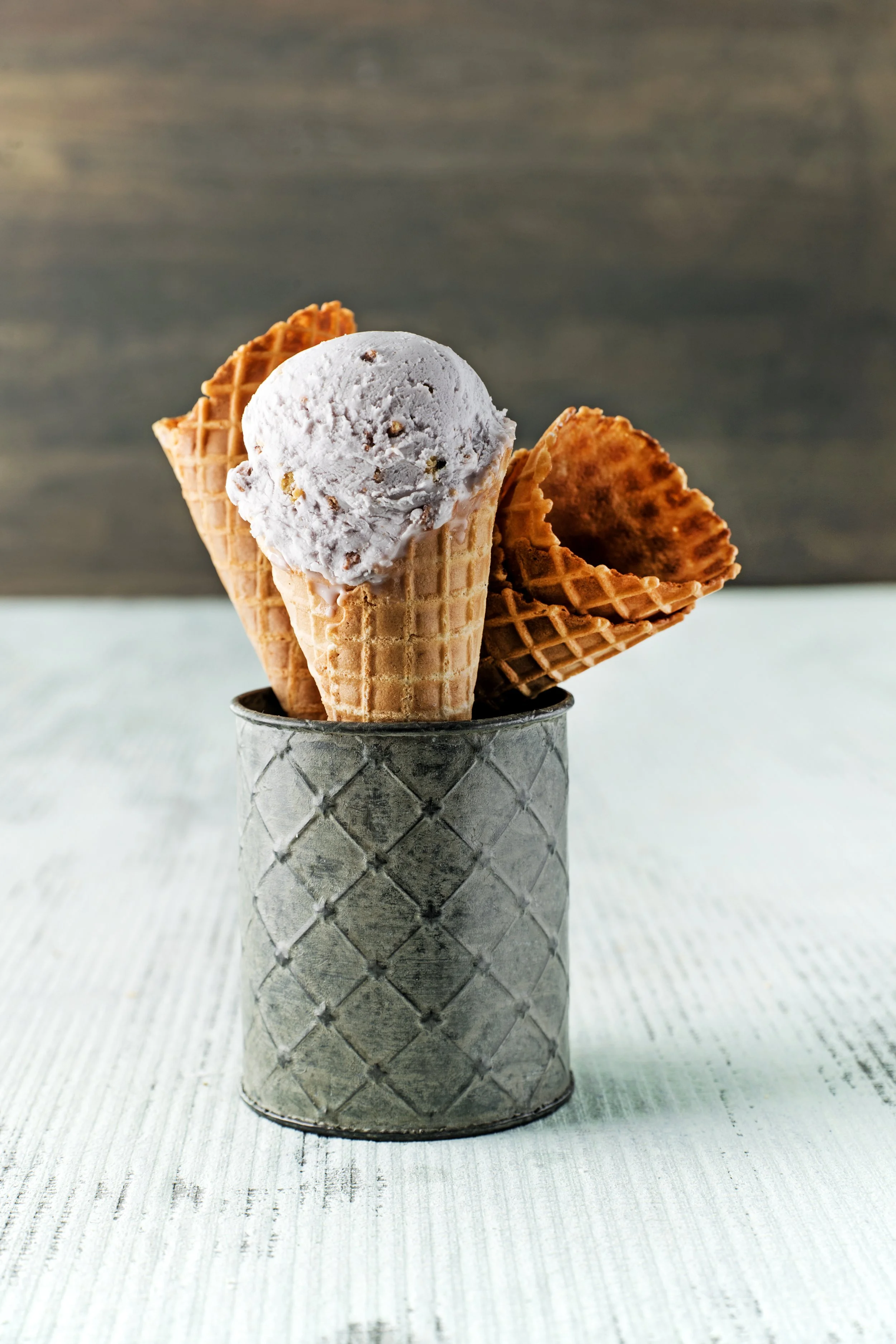 A metal container with a diamond pattern holds three waffle cones, one with a scoop of cookies and cream ice cream and two empty cones.