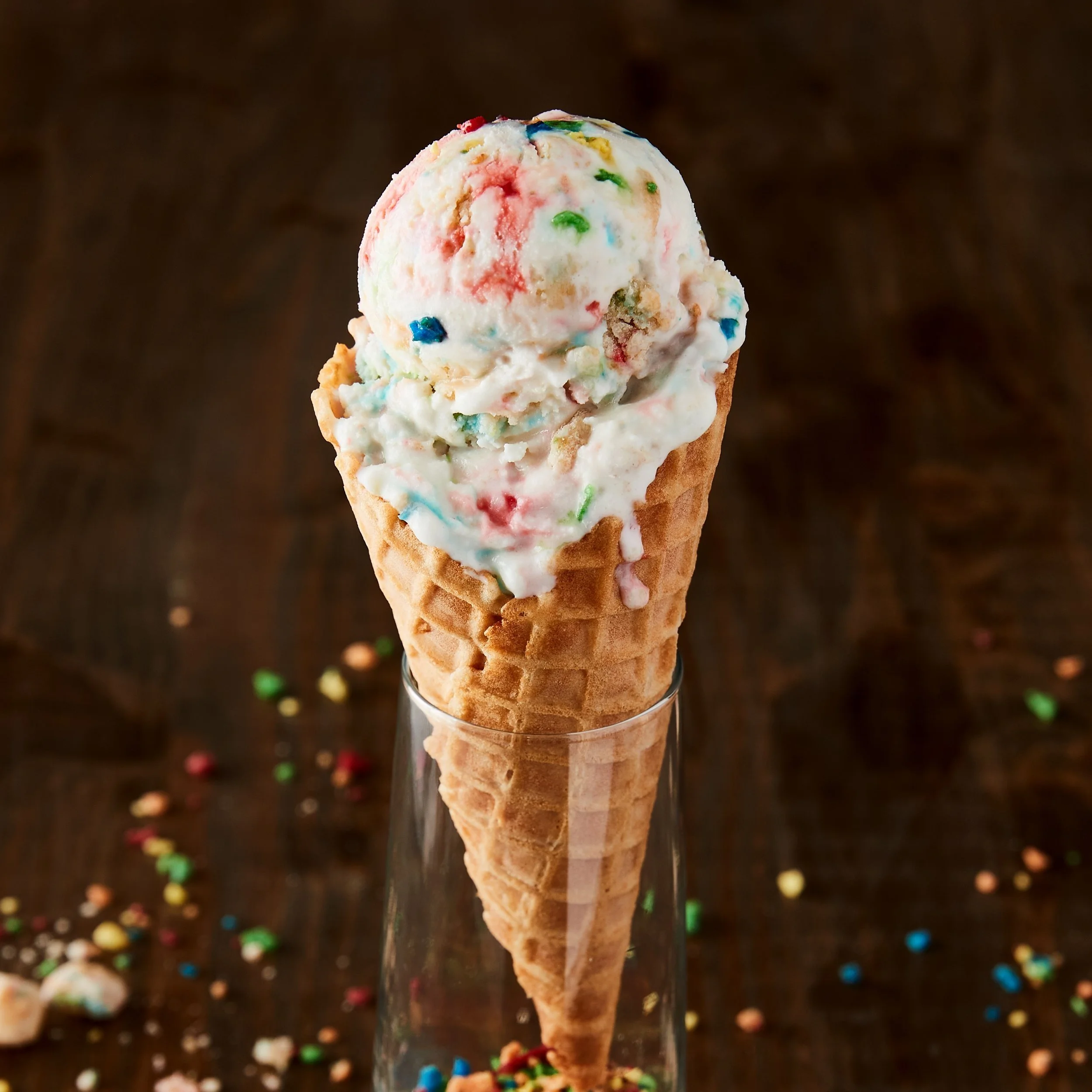 A scoop of colorful birthday cake ice cream in a waffle cone, in a glass container on a dark wooden surface with sprinkles scattered around.