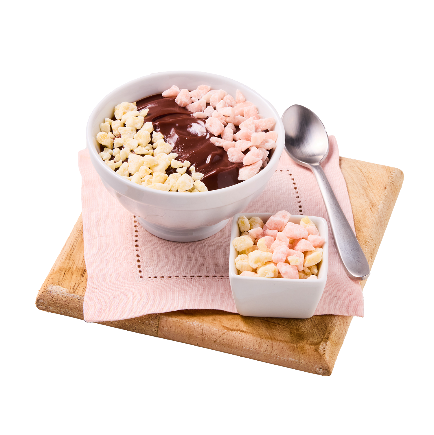 A bowl of chocolate pudding topped with pink and white crunchy cereal pieces, accompanied by a small square dish of the same cereal and a metal spoon, all arranged on a wooden cutting board with a pink cloth underneath.