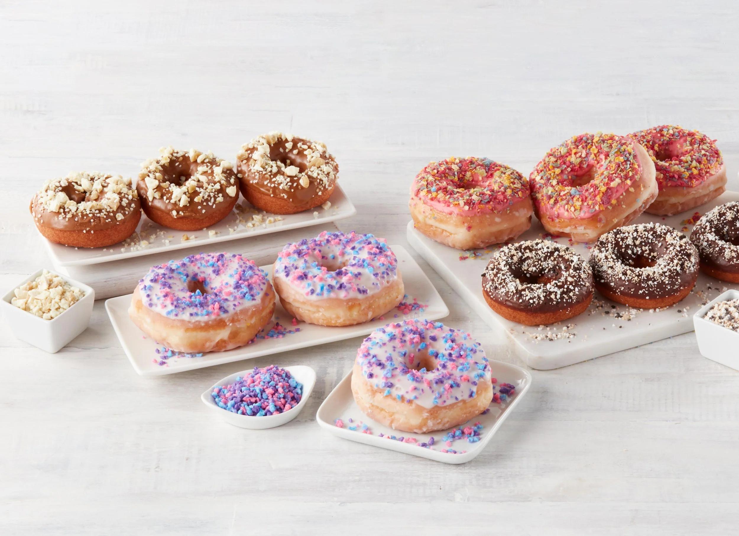 Assorted donuts with colorful sprinkles and toppings on white plates and small bowls, arranged on a white surface.