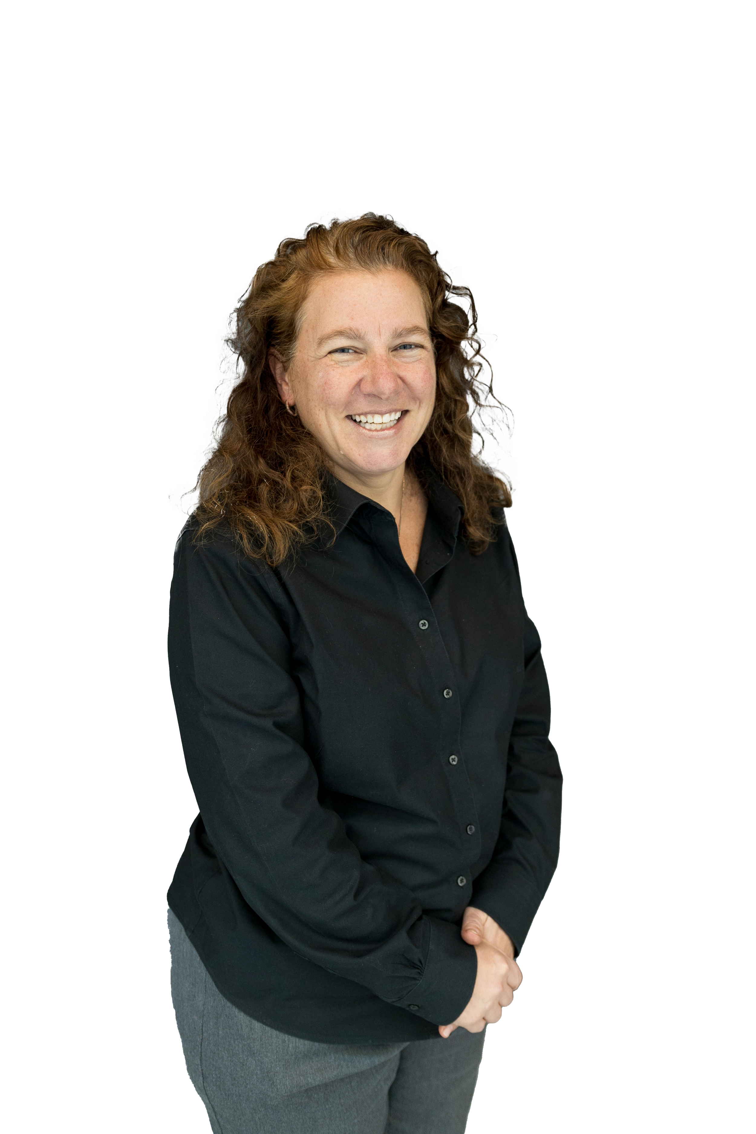A woman with curly red hair, wearing a black button-up shirt and gray pants, standing with her hands clasped in front of her and smiling.