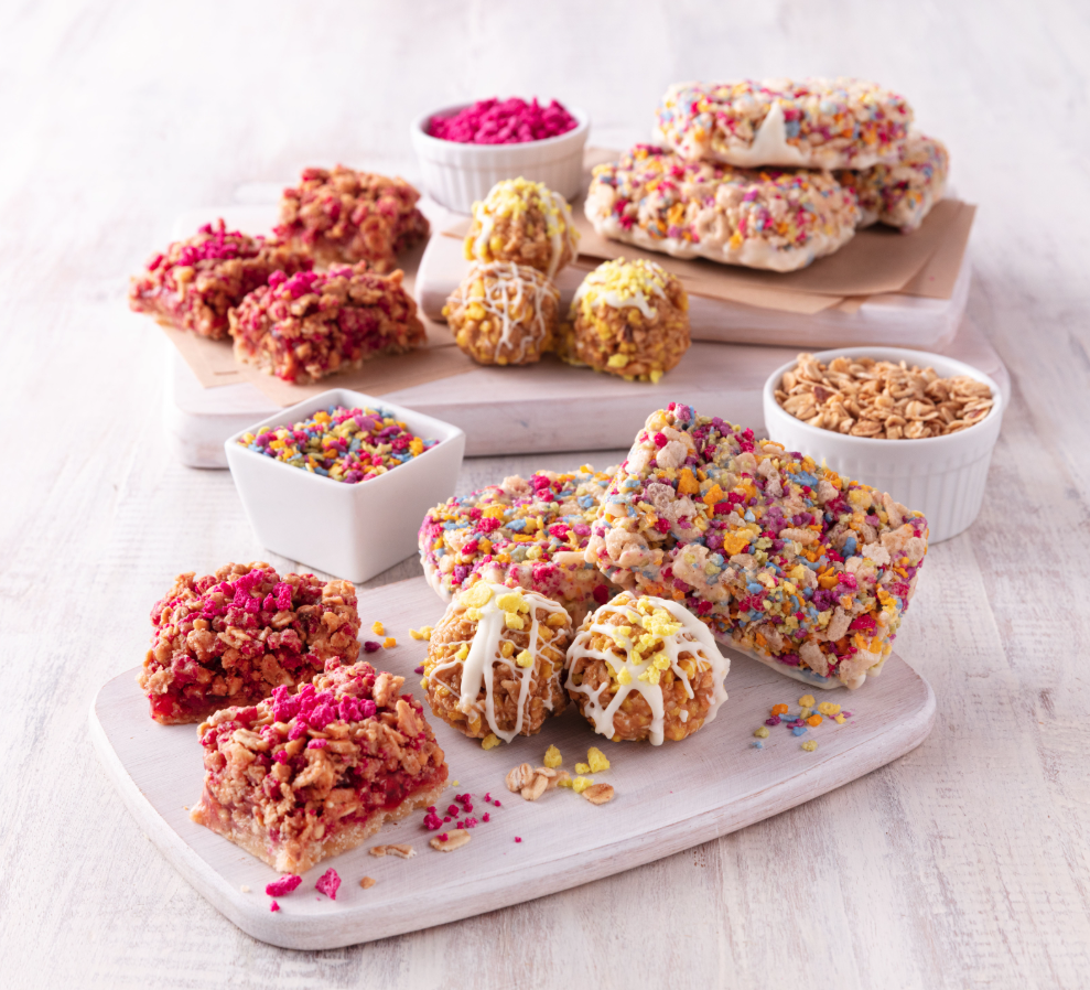Assorted colorful rice krispie treats and candies on white wooden boards with bowls of sprinkles and crushed cereal.