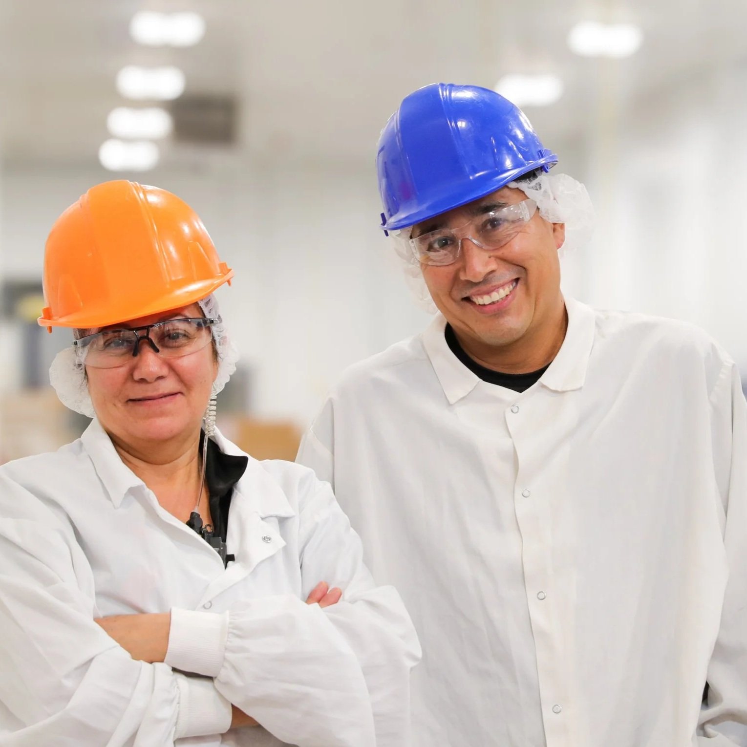 Two factory workers, a woman with an orange safety helmet and glasses, and a man with a blue safety helmet and goggles, smiling in a bright industrial setting.